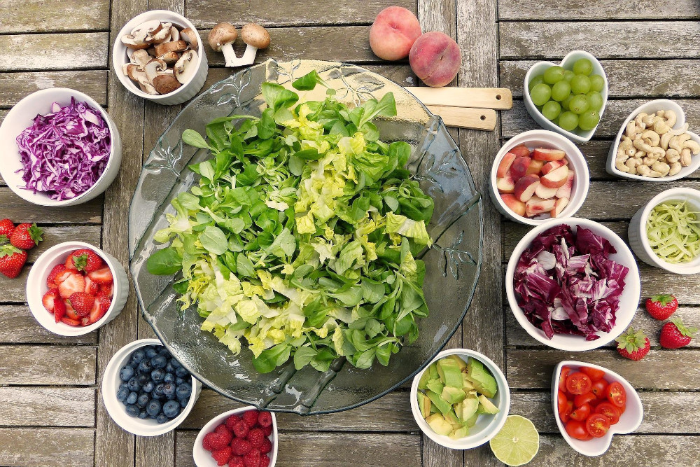 На столі розміщено багато дрібних тарілок в яких знаходяться дрібно порізані продукти харчування: листя салату, полуниця, капуста, томати, гриби, ягоди полуниці, малина, чорниці, горіхи