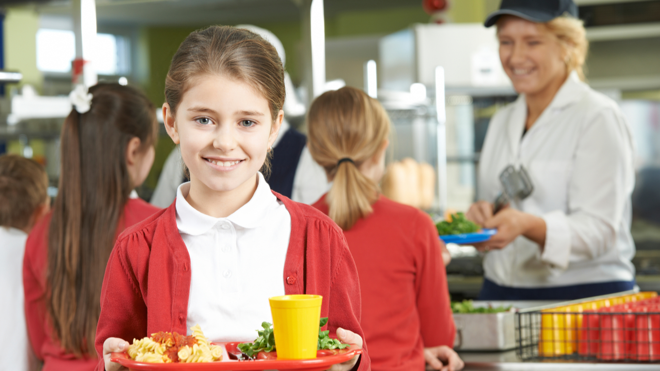 Фото дівчинки, що несе тримає тацю з шкільним обідом. Дівчинка посміхається.